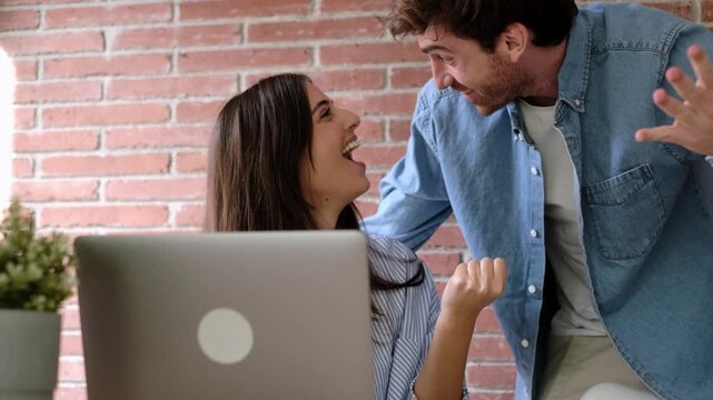 Joyful couple feeling ecstatic while looking at a laptop computer, celebrating a successful achievement, online win, or great news with raised arms and happy expressions at home