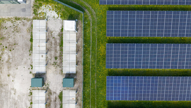 Aerial view of rows of solar panels on a green field with yellow flowers next to concrete foundations and utility infrastructure in Warmenhuizen, North Holland, Netherlands.