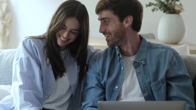Smiling young man and woman sitting together on the couch in their living room, affectionately watching content on a laptop, talking, laughing, and enjoying their shared time at home