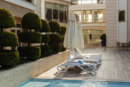 Luxury resort poolside area with white sun loungers and closed umbrellas arranged near clear blue water, modern hotel courtyard with trimmed decorative bushes in warm sunlight, vacation relaxation 