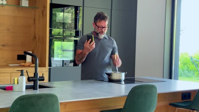 Multitasking man with a beard and glasses talking on the speakerphone of his mobile device while stirring food in a pot on an induction cooktop in a modern kitchen at home
