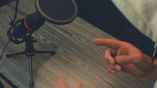 Close up high angle of unidentifiable man in hoodie gesturing while talking propaganda show into a studio microphone. Wooden desk background suitable for podcast recording and vlog content