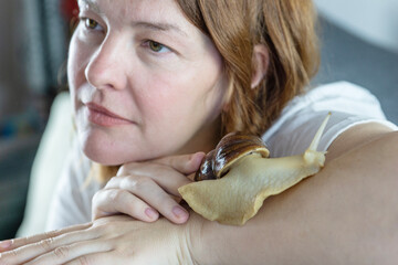 woma with giant achatina snail on her arm looking away with calm expression during beauty treatment in cozy room with natural lighting concept of cosmetology, skincare, wellness © Lana Pietukhova