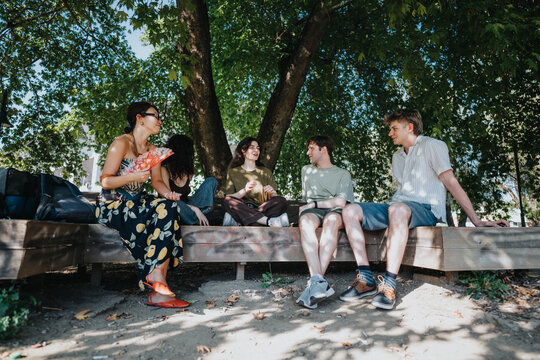 A group of young friends sit and talk on a wooden bench under shady trees. They relax and laugh together in a sunny park setting.