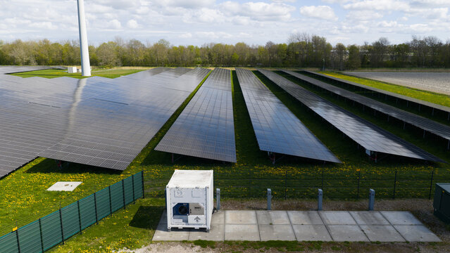 Aerial view of rows of solar panels in a large solar farm with a wind turbine and green fields under a cloudy sky in Warmenhuizen, North Holland, Netherlands.