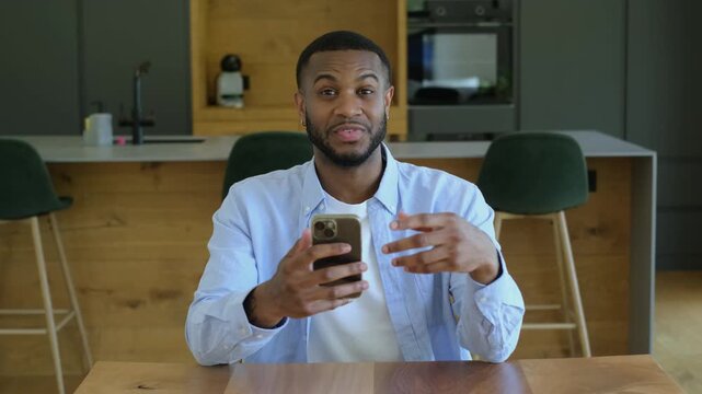 Cheerful black man holding a smartphone and talking during a video call, sitting at a wooden table in his modern kitchen and gesturing with his hands while looking at the camera