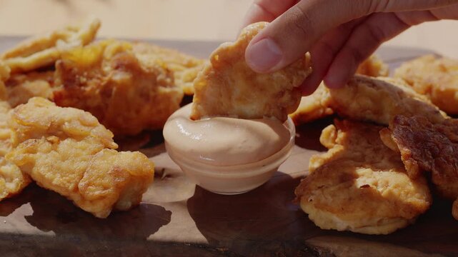 Close-up slow motion footage of a young woman's hand dipping a crispy golden chicken nugget (strip) into a bowl of creamy white sauce (mayonnaise). The video showcases the appetizing texture of the br