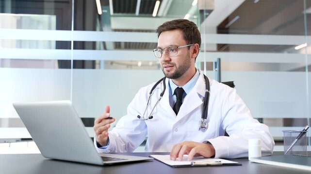 Doctor in white coat talking on video call using laptop sitting at desk in modern hospital office. Professional healthcare worker explaining medical information to patient during online consultation.
