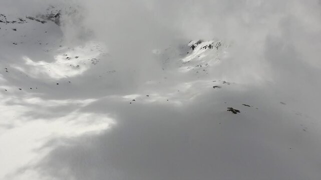 Aerial cinematic view reveals a snowy mountain slope emerging softly through drifting clouds. Winter mountains, alpine terrain, and dramatic sky express solo airsport action in minimal composition.