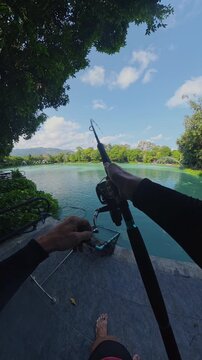 Vertical video. First Person View Fighting Huge Fish With Spinning Rod Near Lake Shore, Line Pulling From Reel Under Strong Tension, Outdoor Pond Setting, Wide Angle Fishing Action
