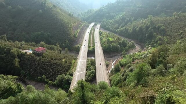 An aerial view shows road bridges and highway tunnels crossing a forested mountain valley landscape. Elevated overpasses and curving underpasses connect transport routes amid dense green hills.