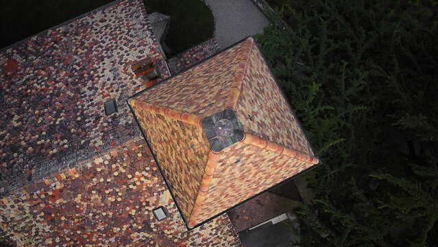 Aerial view of the distinctive multi-colored tiled roof and square tower of a historic building surrounded by green trees in Luins, Vaud, Switzerland.