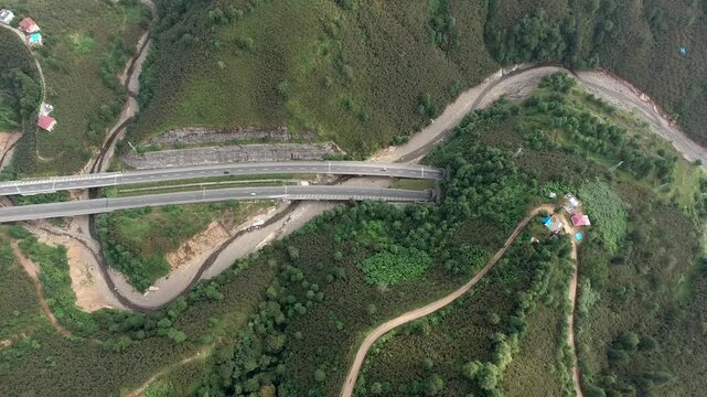 An aerial view shows road bridges and highway tunnels crossing a forested mountain valley landscape. Elevated overpasses and curving underpasses connect transport routes amid dense green hills.