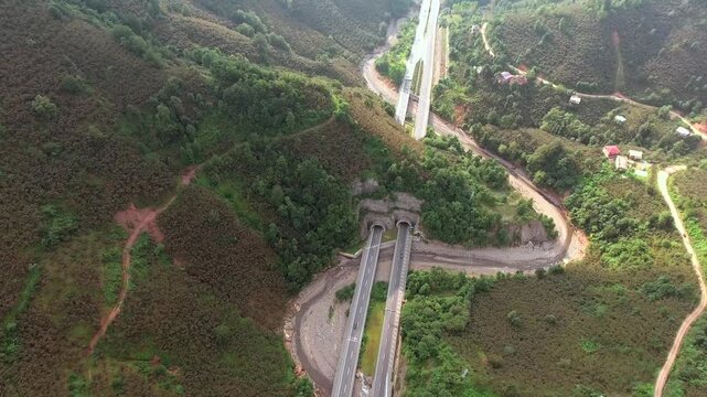 An aerial view shows road bridges and highway tunnels crossing a forested mountain valley landscape. Elevated overpasses and curving underpasses connect transport routes amid dense green hills.