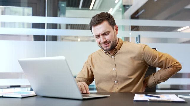 Tired businessman suffering from lower back pain while working on laptop at desk in modern office, massaging sore muscles. Discomfort, strain, poor posture during long hours of sedentary work routine.