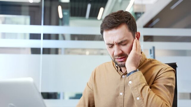 Close up of sick businessman suffering from ear pain while sitting at desk in office, touching ear with fingers in discomfort. Ear infection, otitis media and health issue affecting daily work routine