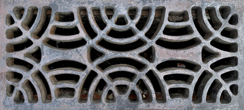 Close up of cast iron drainage grate in rustic weathered grey metal. Symmetrical, Art Deco-inspired geometric pattern with interlocking arcs and straight lines. Seamless background texture