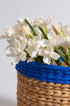 Close-up of woven basket filled with white flowers and green stems against a clean white backdrop.
