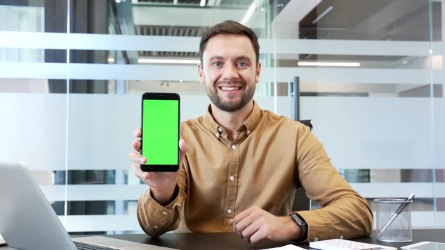 Businessman sitting at desk in modern office holding smartphone with green screen and looking at camera. Mockup template, chroma key display for mobile app advertising, digital presentation corporate