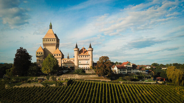 Aerial view of the medieval Vufflens-le-Chateau castle with its prominent square tower and turrets overlooking lush green vineyards under a blue sky in Vufflens-le-Chateau, Vaud, Switzerland.