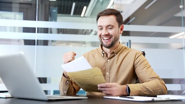 Smiling surprised businessman reading positive letter while sitting at desk in modern office, joy and satisfaction. Great news, success moment and celebration during productive corporate workflow.