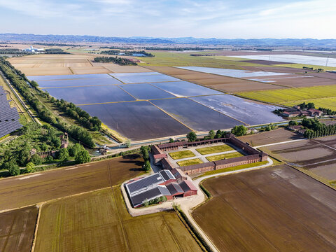 Aerial view of Tenuta Colombara featuring flooded rice fields, traditional farm buildings, and solar panels under a clear sky in Livorno Ferraris, Piemonte, Italy.