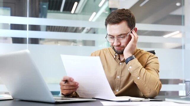Bored businessman holding document and working with paperwork at desk in modern office, showing low motivation, lack of interest. Tired employee feeling disengaged during routine administrative tasks.