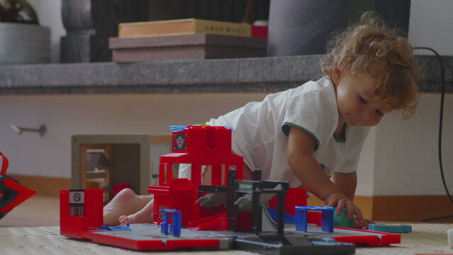 Toddler kneeling on floor reaching for toy garage pieces, curious and engaged, using hands to build and move cars, creative play and problem-solving during quiet childhood moment