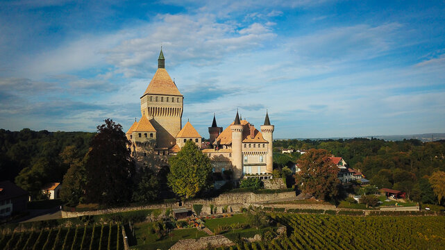 Aerial view of the medieval Vufflens Castle with its prominent square keep and turrets surrounded by lush vineyards and green trees under a blue sky in Vufflens-le-Chateau, Vaud, Switzerland.