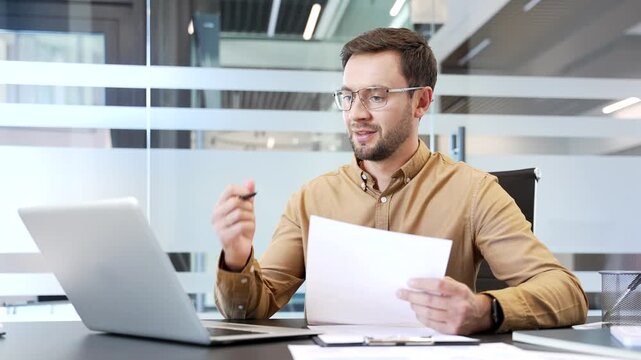 Smiling businessman reviewing financial documents and looking at laptop screen in office, satisfied with results. Positive outcome, successful analysis and confident business corporate performance