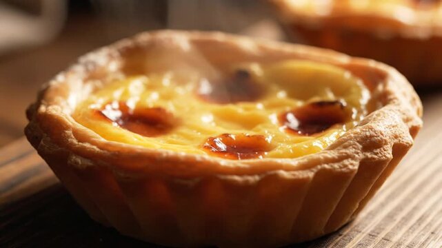 Close-up of a freshly baked Portuguese egg tart featuring a flaky golden crust and caramelized custard top on a rustic wooden surface with steam rising from the warm pastry.