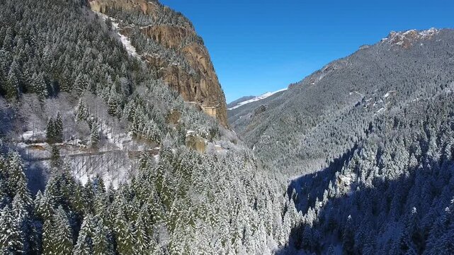 Aerial drone view captures snow covered Sumela Monastery clinging to steep rocky mountain cliff. Forested valley shows valley slope, winter season, and ancient christian plus christian site.