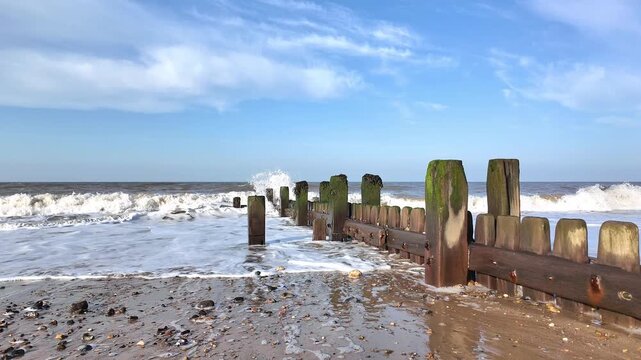 Waves crash against weathered wooden groynes on the Norfolk coast, sending white spray into the air as the tide rolls in beneath a windswept sky.