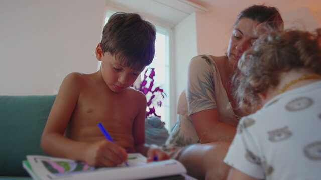 Boy focused on home activity while writing in book with blue marker, mother supervising closely and toddler playing nearby, calm family learning moment in cozy living room