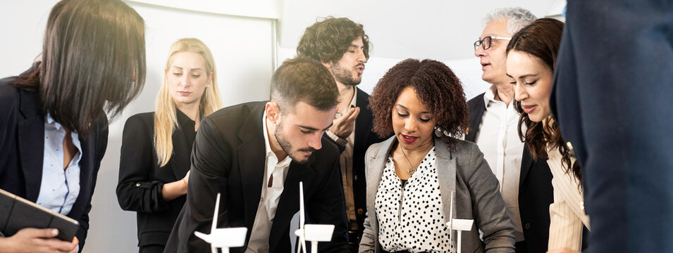 Multicultural team of engineers and business professionals discussing wind turbine models and renewable energy strategy in modern office meeting. Millennials and generation x colleagues