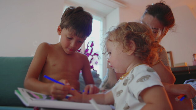 Boy focused on home activity while writing in book with blue marker, mother supervising closely and toddler playing nearby, calm family learning moment in cozy living room