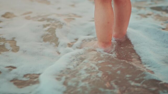 Close-up of an Asian toddler's feet standing on a sandy beach while white sea foam washes over them at sunset. Childhood discovery and sensory play during a family vacation in warm.