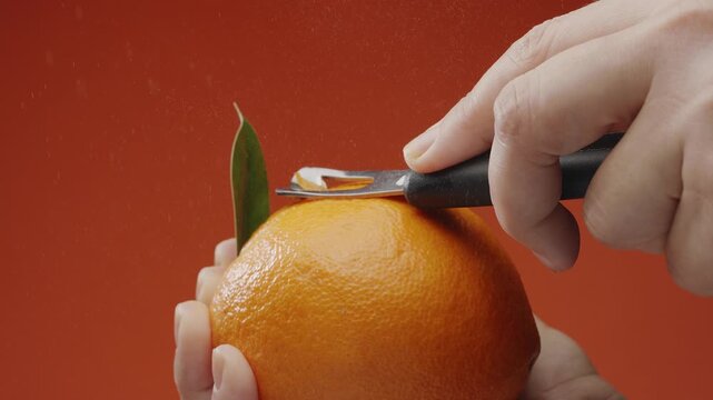 Getting orange peel with a special kitchen tool; orange juice with essential oils sprinkling out under the cut on orange background. Slow motion macro video. 