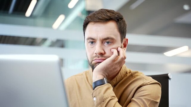Close up of bored businessman sitting at desk in office, leaning chin on hand, looking at laptop screen with uninterested expression. Low motivation, routine fatigue and lack of engagement at work.