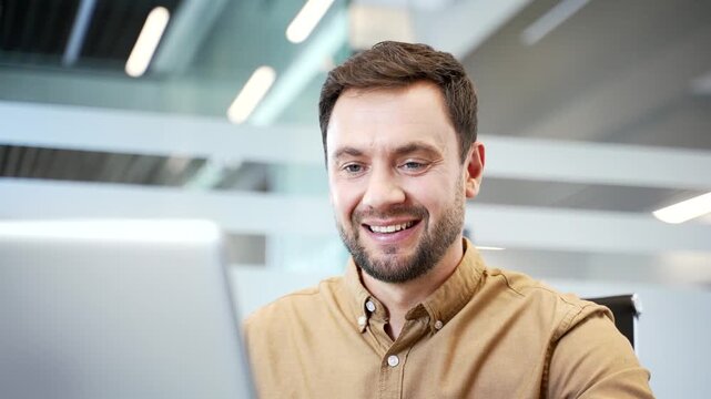Close up of businessman reading positive message on laptop at desk in office, smiling as mood shifts to joy. Genuine reaction to good news, pleasant surprise and uplifted spirit during work routine.