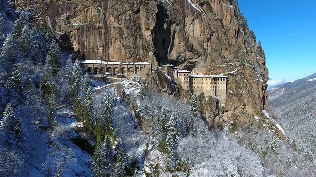 Aerial drone view captures snow covered Sumela Monastery clinging to steep rocky mountain cliff. Forested valley shows valley slope, winter season, and ancient christian plus christian site.