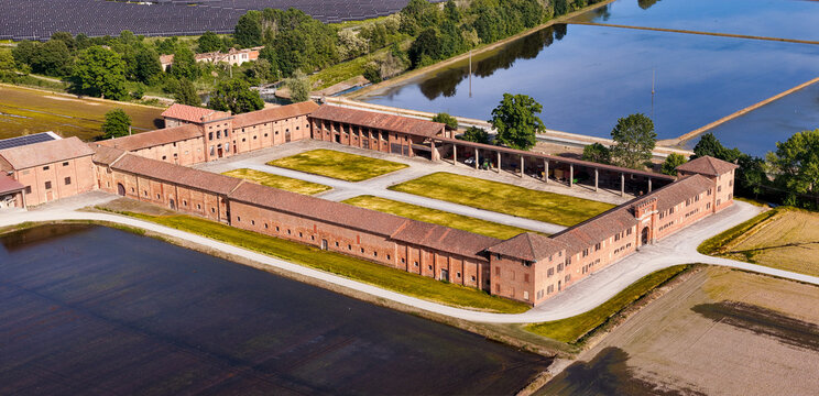Aerial view of Tenuta Colombara, a historic brick farmstead with a large courtyard surrounded by flooded rice paddies and green trees in Livorno Ferraris, Piemonte, Italy.