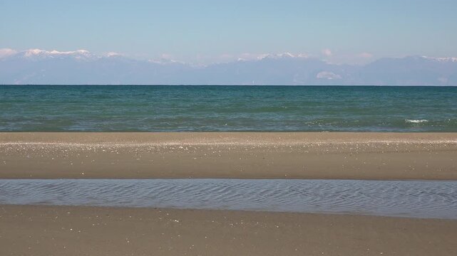 Longshore drift shapes tombolo and sand bar beside tranquil beach near open sea shore. Coastal sediment movement forms sandy barrier with gentle shoreline curves beneath clear sky.