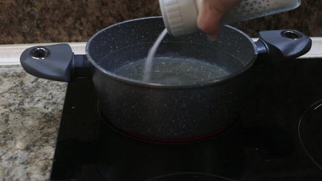 salt being poured into boiling water in black granite pot on induction stove kitchen counter preparation before cooking dumplings