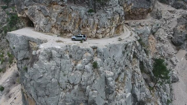 Aerial view of car driving along extremely dangerous rocky mountain cliff road track. Rugged vehicle follows a perilous ledge route above steep stone slopes in remote Himalaya.