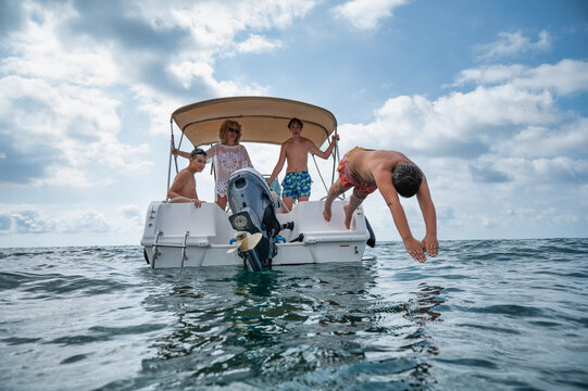 Middle aged mother with her three teenage sons on a motorboat during summer vacation, watching one boy dive into the sea in a joyful family adventure moment