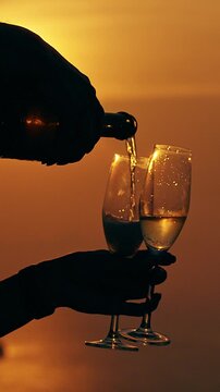 Vertical video. Close View of Champagne Pouring into Glasses in Hands Silhouette against Golden Sunset Ocean Warm Light Celebration Romantic Leisure Scene
