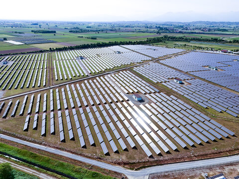 Aerial view of a large solar farm with rows of photovoltaic panels reflecting sunlight under a clear sky in Torrazza Piemonte, Piemonte, Italy.
