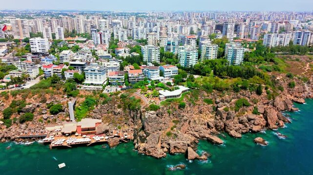Coastal cliff with mostly low/mid-rise buildings near the edge; a large high-rise skyline in the background, near Lower Düden Waterfalls, Antalya, Turkey.