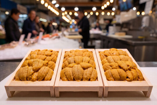 Fresh yellow sea urchin roe displayed in three small wooden trays at a busy indoor seafood market with blurred customers and warm lights in the background.